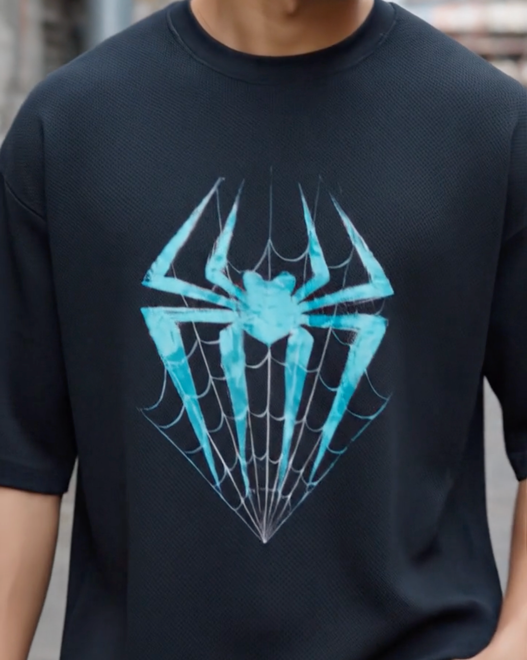 Front view of a black oversized t-shirt worn by a model from Lowkey fashion featuring a teal spider and web graphic in the center.