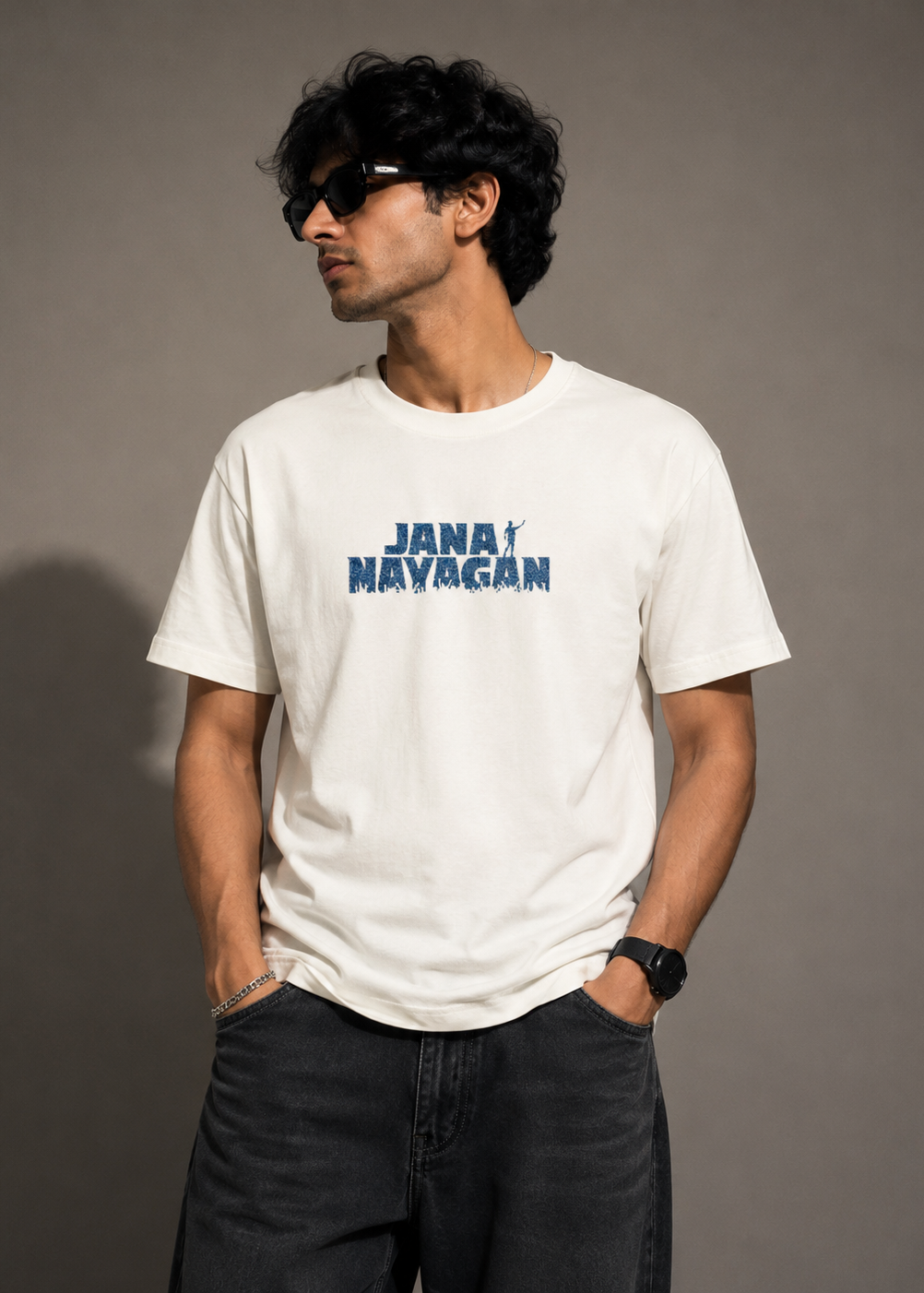A man wearing a white crewneck t-shirt with a blue 'JANA NAYAGAN' graphic on the chest, paired with dark gray jeans, set against a plain gray studio background.