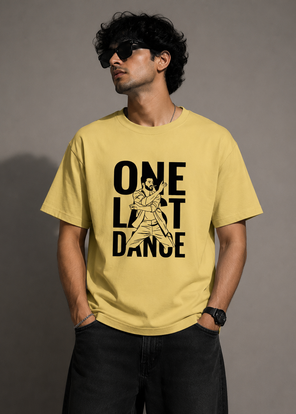 A man wearing a pale yellow graphic t-shirt with the text 'ONE LAST DANCE' and a dancer illustration in black, paired with black jeans against a neutral gray studio background.
