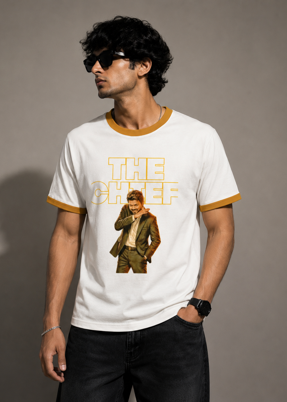 A man wearing a white ringer t-shirt with mustard yellow trim and a graphic print of 'The Chef' on the front, paired with black jeans against a neutral gray studio background.