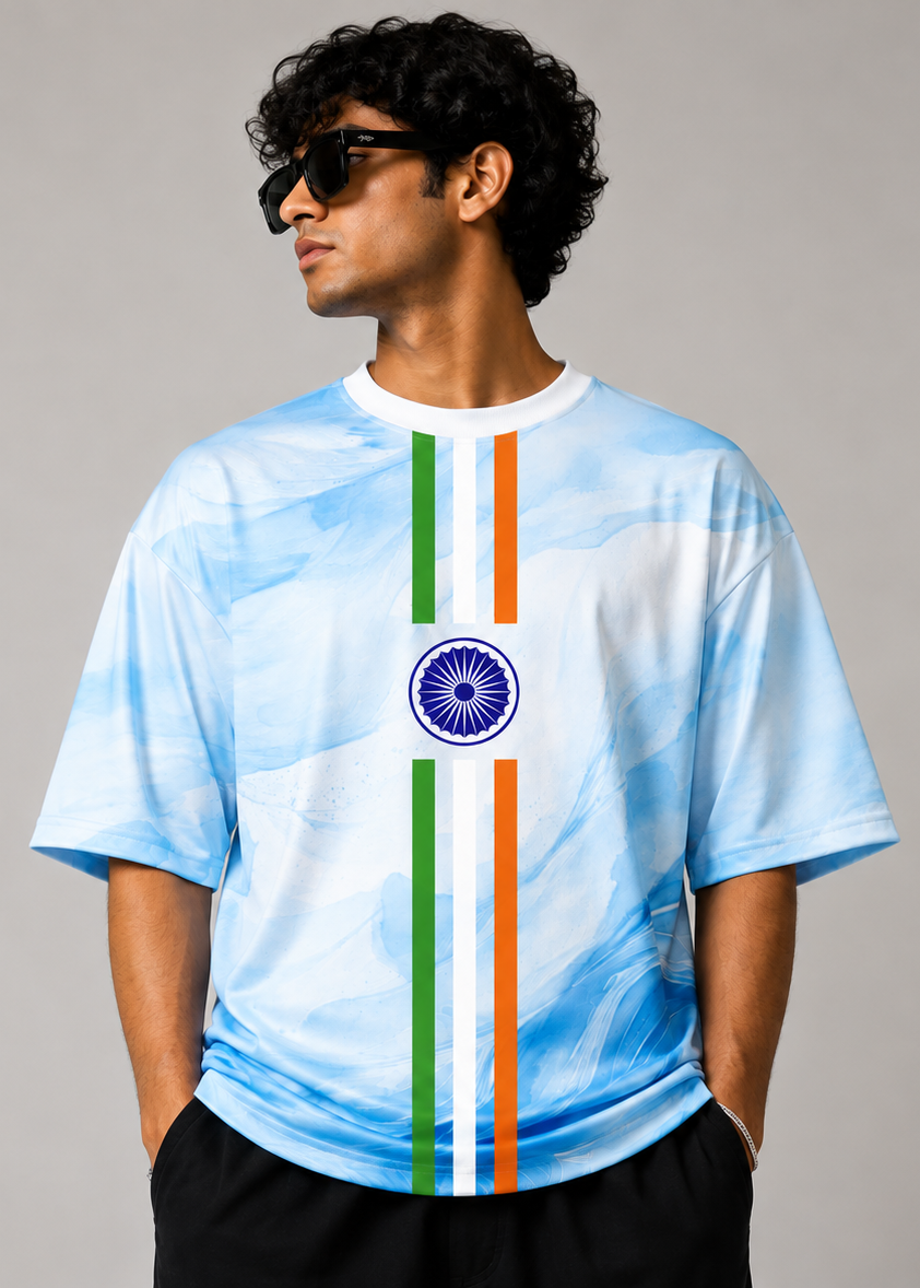 A man wearing a light blue and white watercolor-patterned t-shirt with a vertical Indian tricolor stripe and the Ashoka Chakra emblem on the chest, paired with black shorts and sunglasses, set against a plain gray studio background.